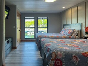 A bedroom with two beds, a TV mounted on the wall, and a balcony with a view of greenery and water.