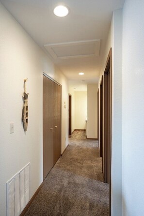 Un couloir recouvert de moquette, avec une porte en bois et une grille d’aération sur le mur.
