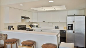 Una cocina con armarios blancos, un refrigerador de acero inoxidable, una cafetera, un tostador y una estufa. Hay tres taburetes de madera en la encimera.