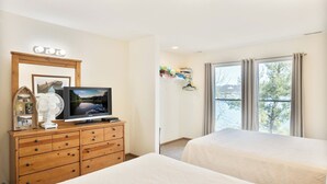 Habitación de hotel con dos camas, un televisor sobre una cómoda de madera, un ventanal con cortinas y vistas a árboles.