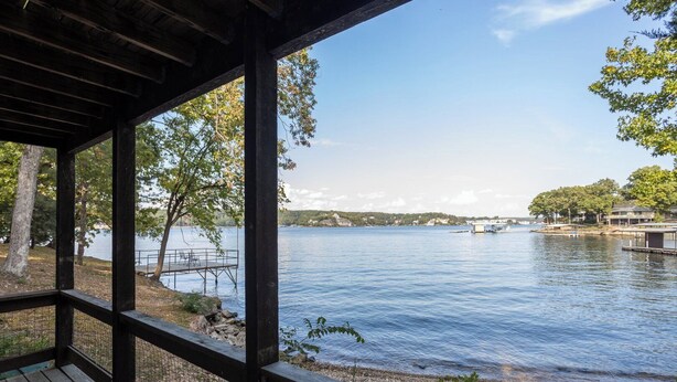 Terraza de madera con vistas a un lago tranquilo, con un muelle y sillas, rodeada de árboles y edificios a lo lejos.