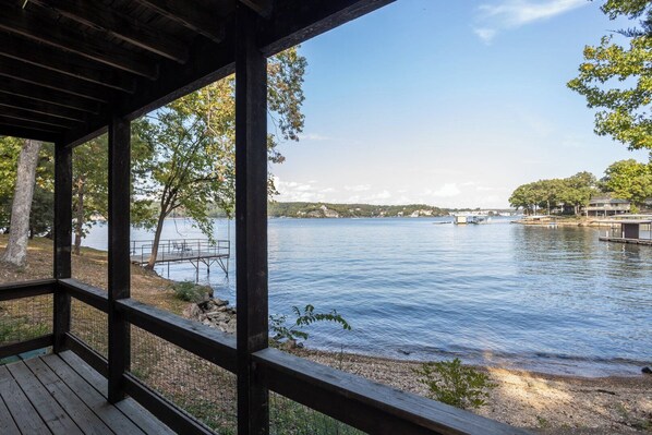 A wooden deck overlooking a calm lake with a dock and chairs, surrounded by trees and buildings in the distance.