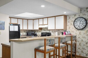 Una cocina con una isla central, taburetes de bar, un refrigerador, un microondas y un reloj en la pared.