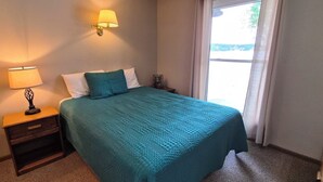 A hotel room with a bed, a nightstand with a lamp, a wall-mounted lamp, and a window with curtains.