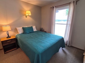 A hotel room with a bed, a nightstand with a lamp, a wall-mounted lamp, and a window with curtains.