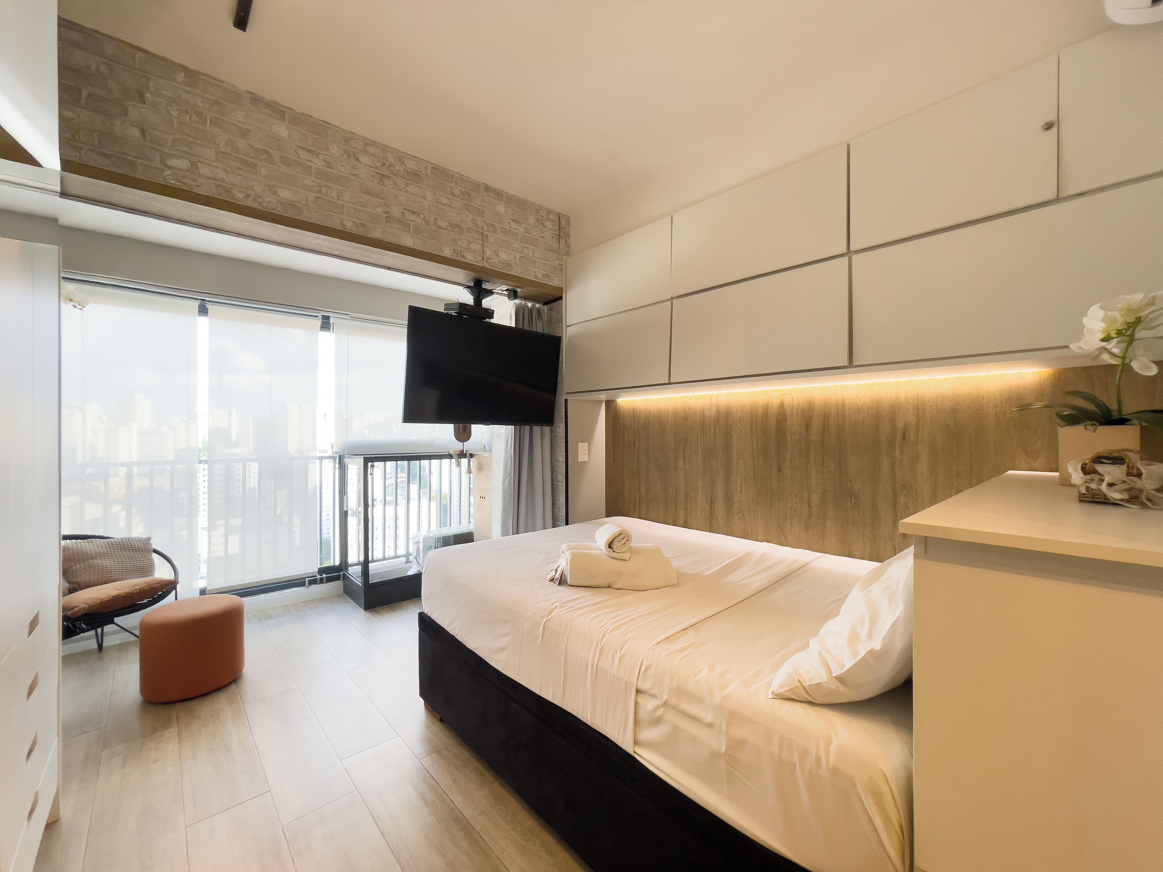 Een moderne hotelkamer met een groot bed, een flatscreen televisie aan de muur, een balkon met uitzicht over de stad en een houten hoofdbord.