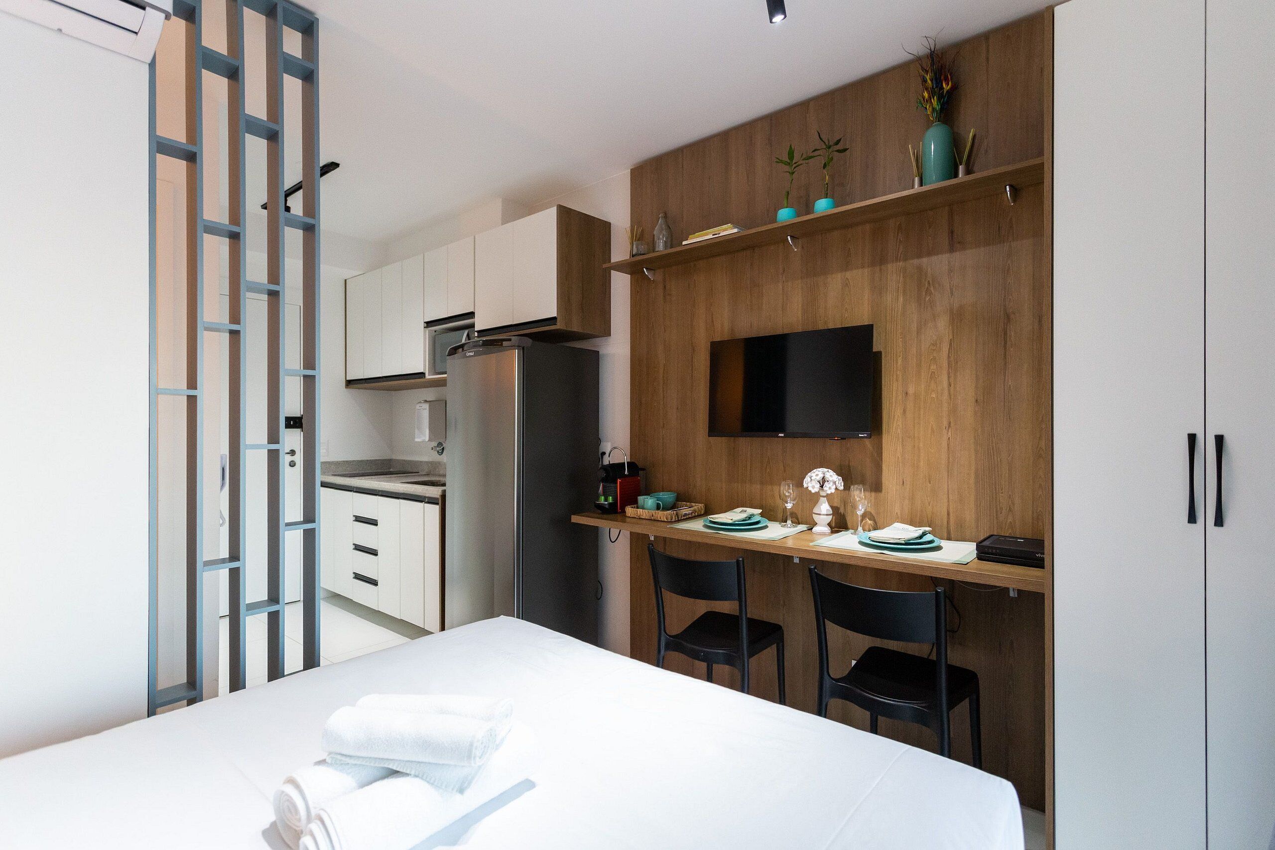 A modern bedroom with a bed, a desk with chairs, a TV, and a wardrobe.