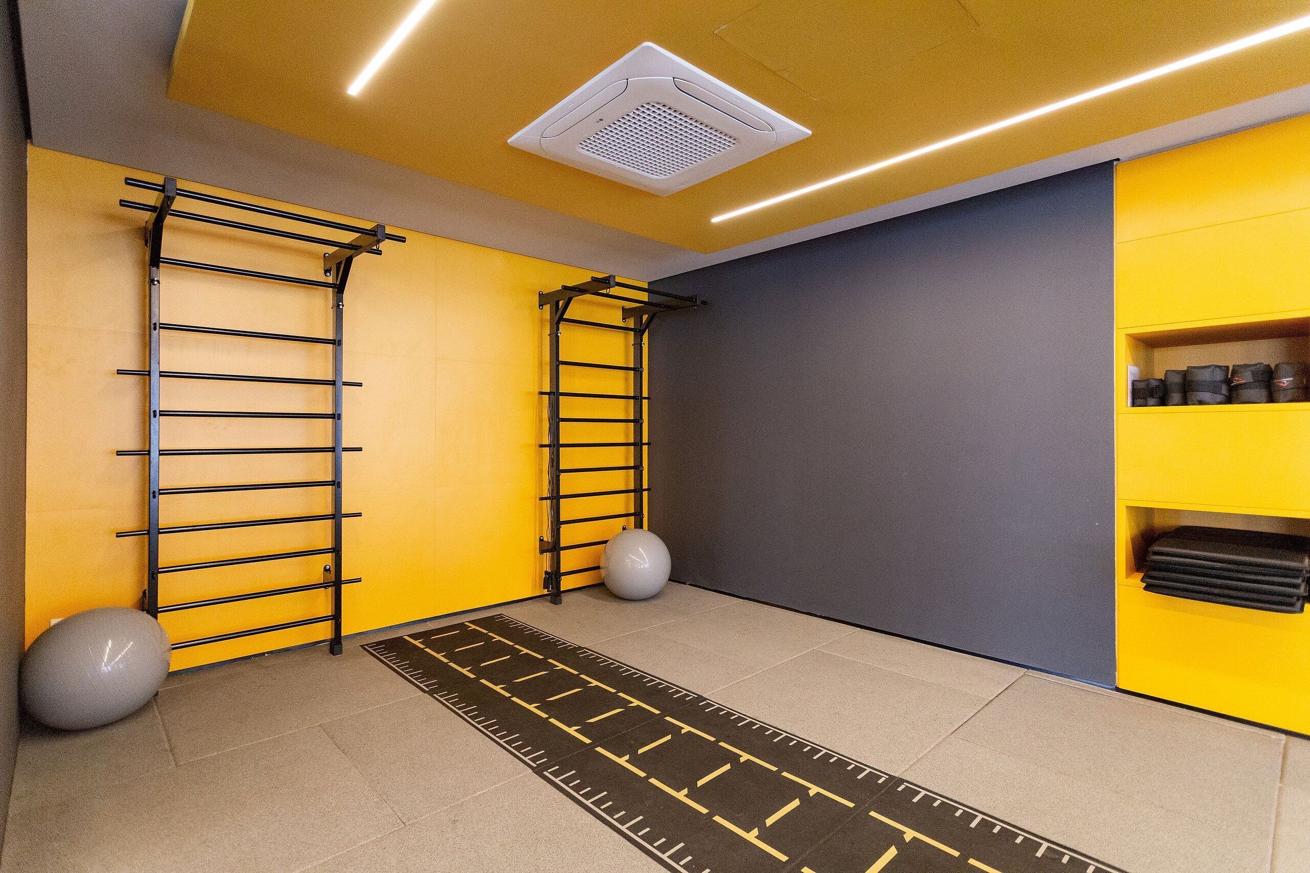 Ein Zimmer mit gelber Wand, grauer Wand und einer schwarzen Stange zum Turnen. Es gibt einen weißen Deckenlüfter und eine schwarze Matte auf dem Boden.