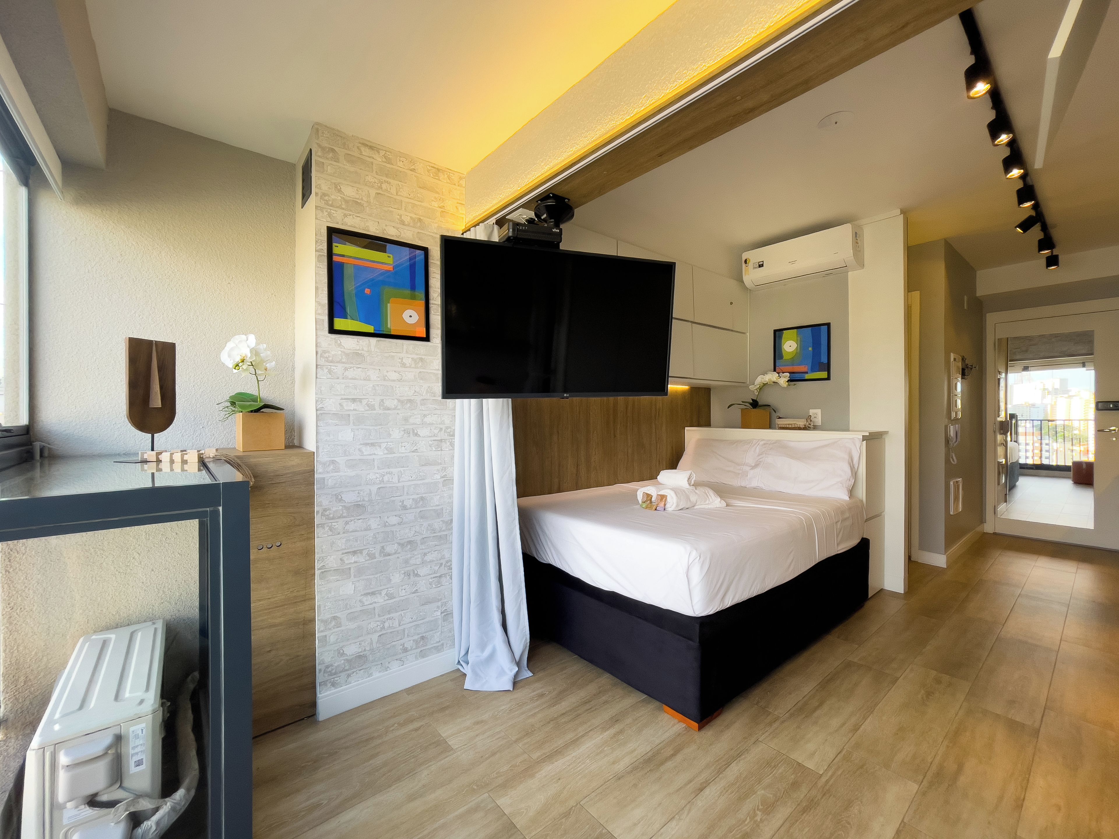 Habitación de hotel moderna con cama, televisión de pantalla plana, escritorio con estructura de madera y vista a la ciudad desde el balcón.