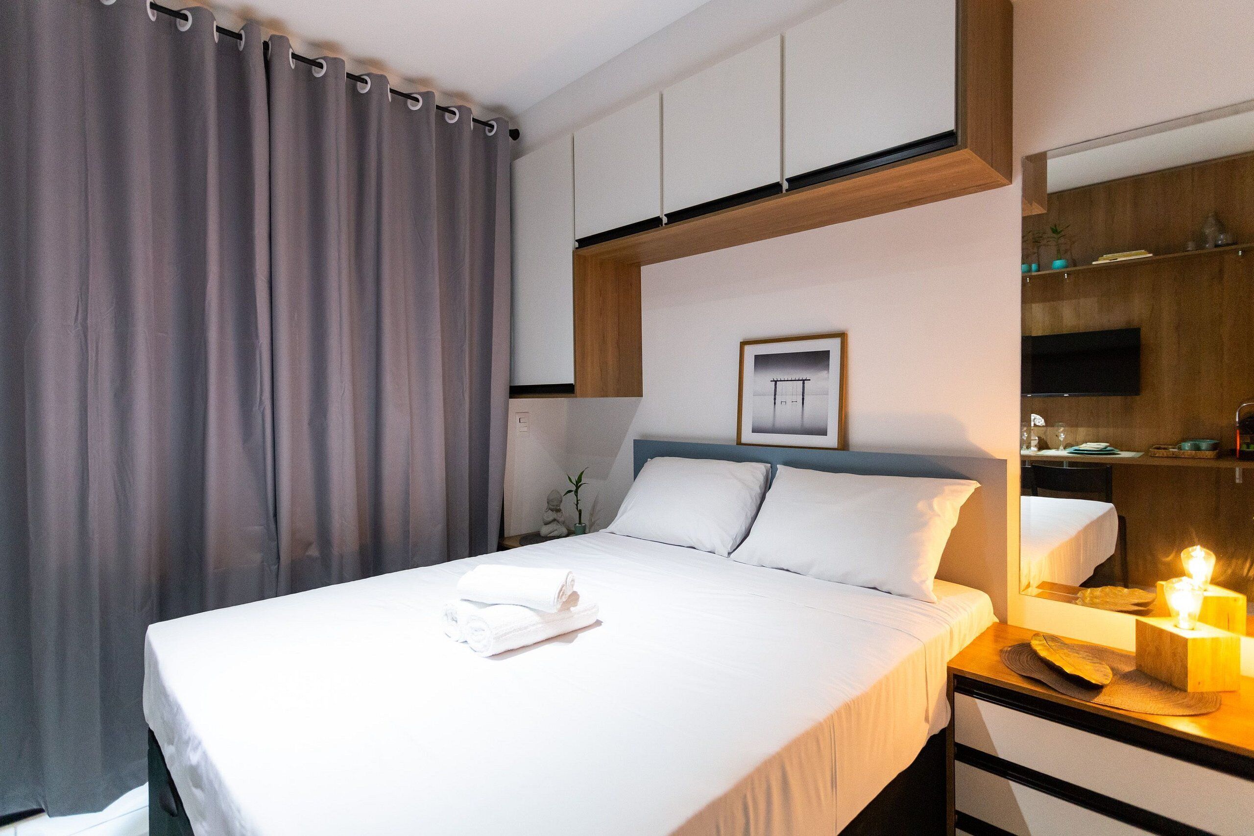 A neatly made bed with white linens, a bedside table with a lamp, and a large window with curtains.