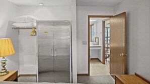A hotel room with a closed wardrobe, a bedside table with a lamp, a small window, and a view of the bathroom.