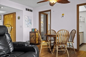 A living room with a black leather sofa, wooden dining table with chairs, a wooden cabinet with decorative items, a ceiling fan, and a map on the wall.