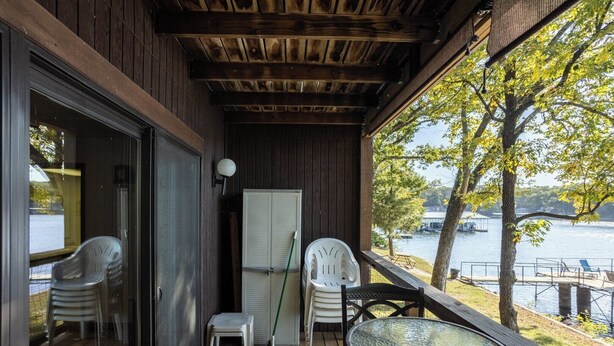 A wooden deck with a glass door leading to a room, a small table with chairs, a broom, and a window overlooking a lake.
