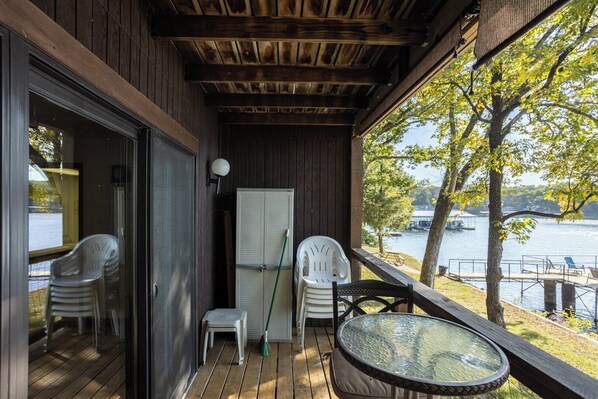 A wooden deck with a glass door leading to a room, a small table with chairs, a broom, and a window overlooking a lake.