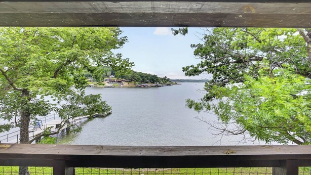 Vista de un lago desde una terraza de madera con barandilla de metal, rodeada de árboles y una orilla distante con edificios.