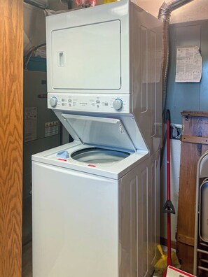 A front-loading washer and dryer unit with a control panel, a vent hose, and a mop leaning against the wall.