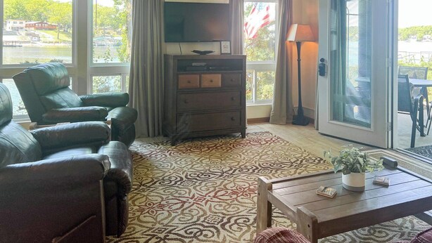 A living room with a sofa, a TV, a wooden cabinet, a patterned rug, and a view of a lake through large windows.
