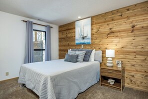 A bedroom with a bed, a nightstand with a lamp, a wooden wall panel, and a window with curtains.