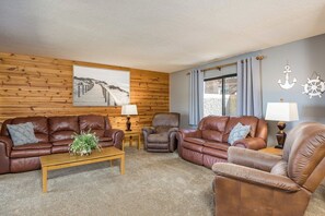A living room with a wooden wall, a large picture, two leather sofas, a coffee table, and a window with curtains.
