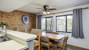 Une cuisine moderne avec un mur en bois, une horloge, une table à manger avec des chaises, un ventilateur de plafond et de grandes fenêtres avec des rideaux.