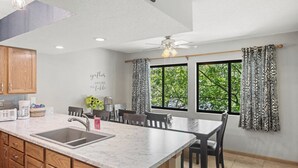 Une cuisine dotée d’un plan de travail en marbre, de meubles en bois, d’un coin repas avec une table et des chaises, d’un ventilateur de plafond et de grandes fenêtres avec des rideaux.