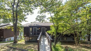 A two-story building with a wooden deck leading to the entrance, surrounded by trees and greenery.