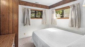 A bedroom with a white brick wall, wooden beams, a large bed, two windows with curtains, and a wooden cabinet.