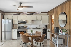 Cocina compacta con armarios blancos, refrigerador de acero inoxidable, microondas, horno y cafetera. Cuenta con una mesa de comedor con sillas y un ventilador de techo.