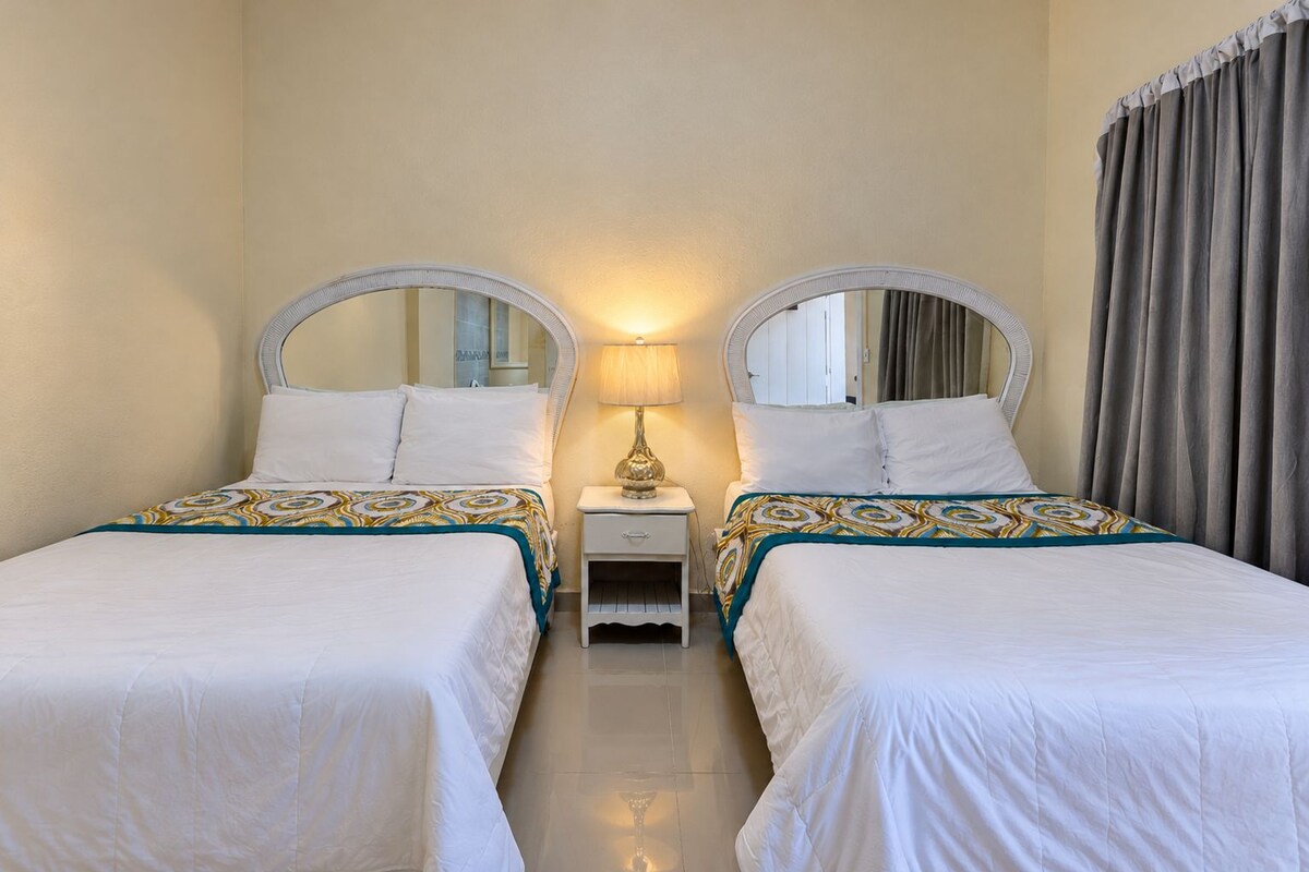 A hotel room with two single beds, a nightstand with a lamp, a mirror, and a window with curtains.