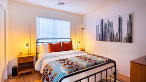 Un dormitorio ordenado con cama, mesitas de noche, una ventana con persianas, una lámpara y un cuadro de un paisaje urbano en la pared.