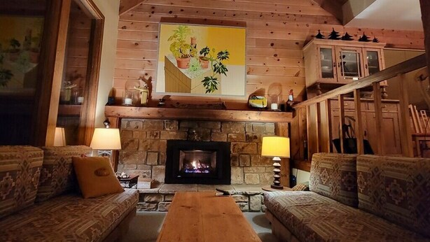 Interior acogedor de una cabaña de madera con chimenea de piedra, muebles de madera y un cuadro sobre la chimenea.