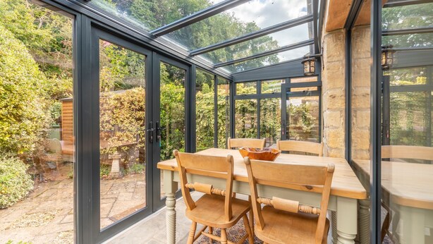 Une véranda avec une table et des chaises en bois, de grandes portes vitrées et une vue sur un jardin.