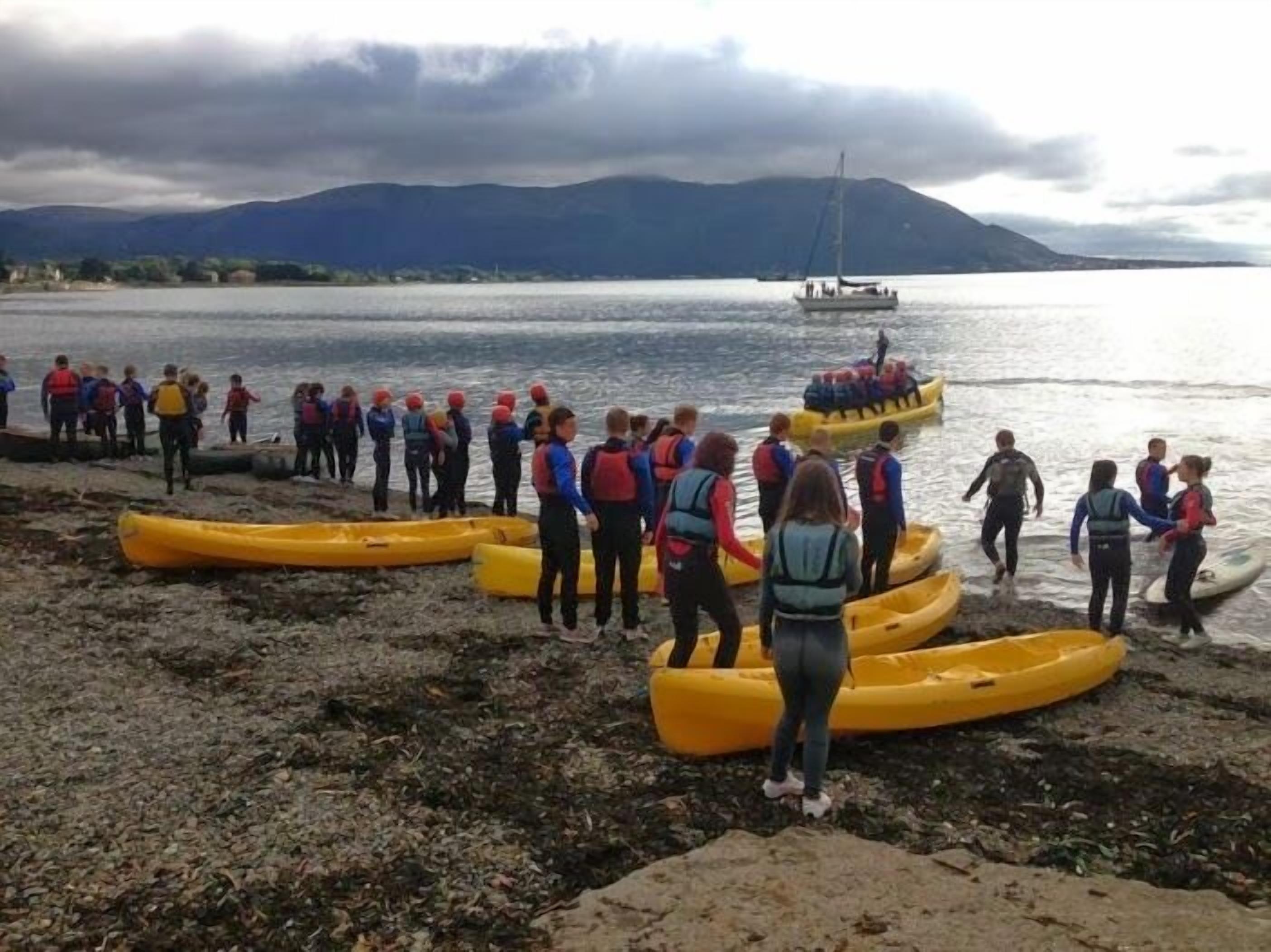 A group of people in yellow kayaks on a rocky shore by a body of water with mountains in the background.
