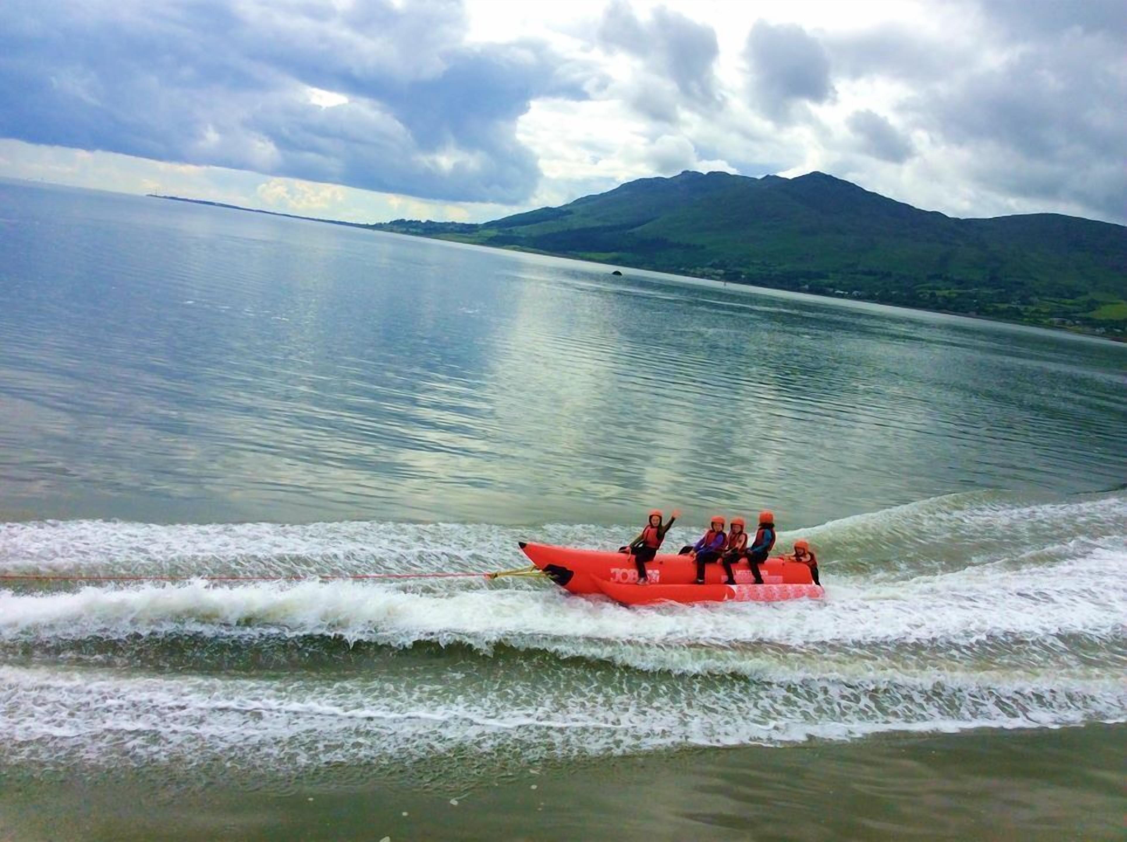 A group of people on a red watercraft moving across a calm lake with mountains in the background.
