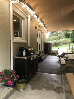 An outdoor covered patio area with a barbecue grill, potted plants, and a wooden table.
