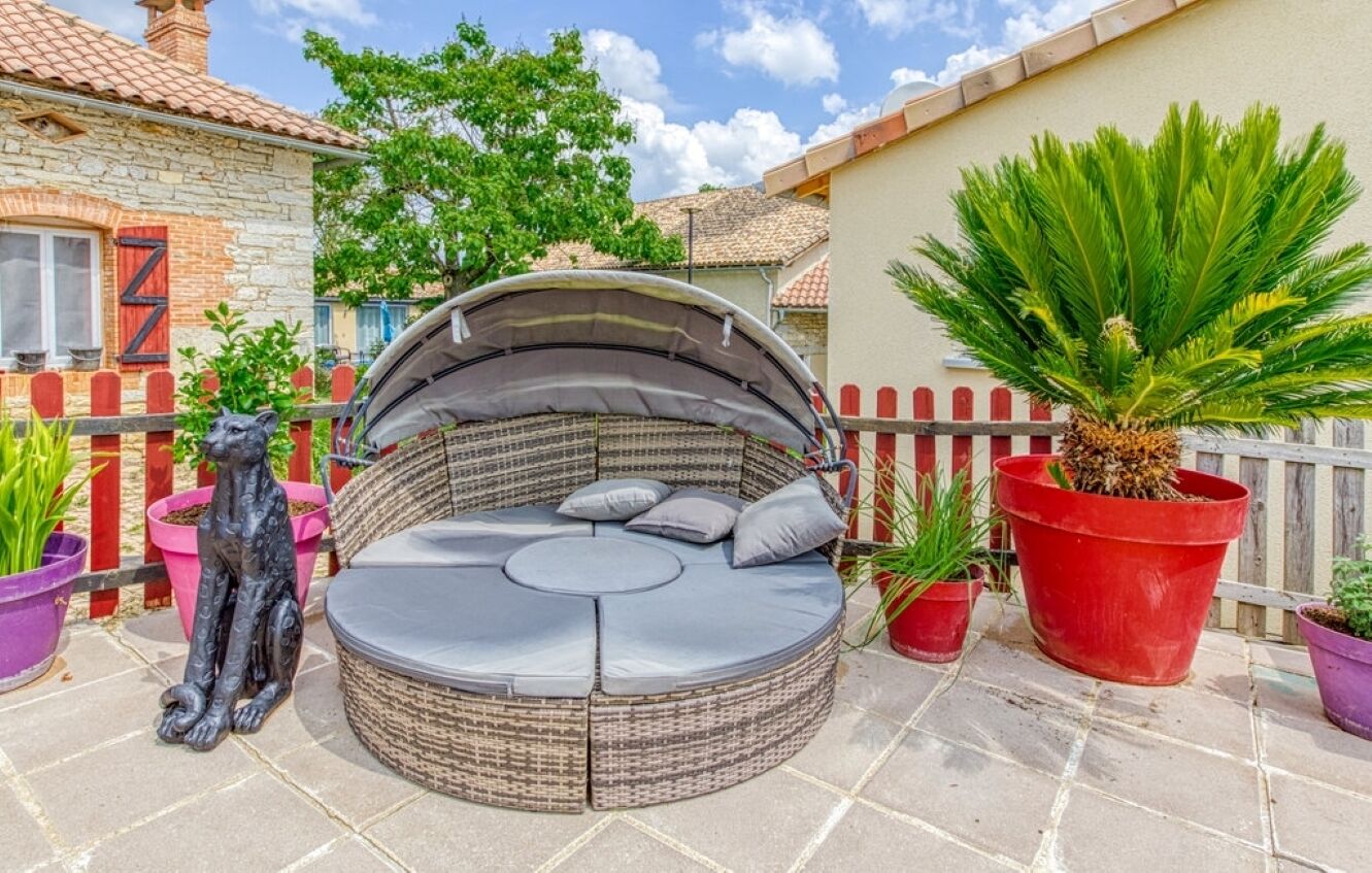 A round wicker sofa with cushions and a canopy, surrounded by potted plants and a decorative statue.