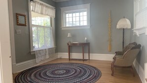 Una habitación con piso de madera, un tapete estampado, una silla, una mesa con lámpara y una ventana con cortinas.