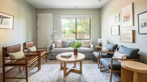 A cozy living room with a sofa, two armchairs, a coffee table, and a side table. There is a door, a window with blinds, and framed pictures on the wall.