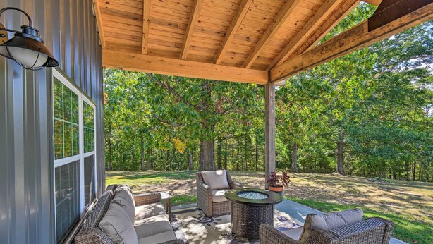 A covered patio with wicker furniture, a fire pit, and a view of a wooded area. - Cozy Tennessee Country Retreat on 5 Acres! (Pegram)