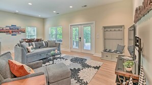 A spacious living room with a grey sofa, a coffee table, and a large floral rug. There are two windows with blinds, a door leading outside, and a staircase with a wooden railing. - Cozy Tennessee Country Retreat on 5 Acres! (Pegram)