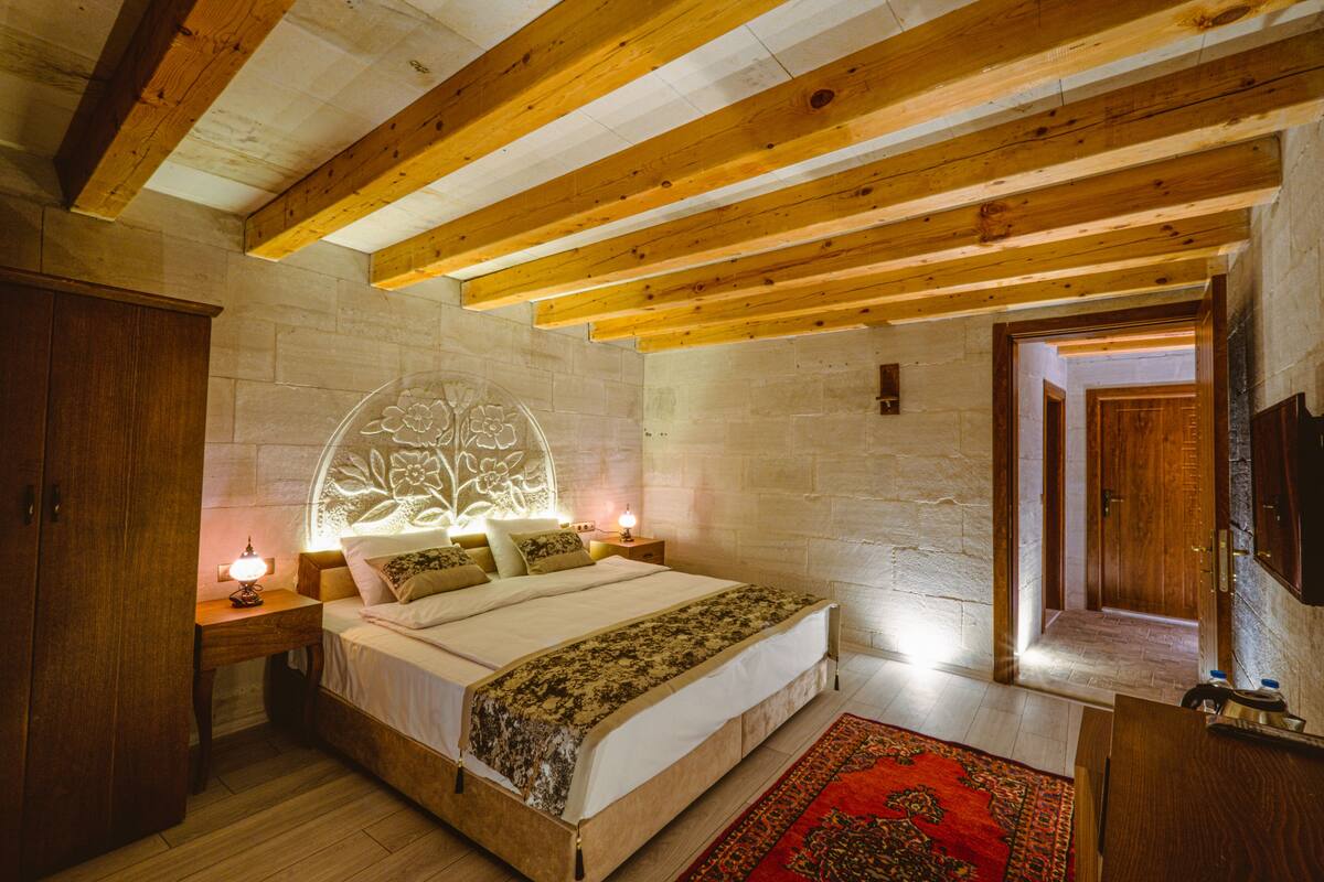 A bedroom with a wooden ceiling, a stone wall, a bed with a floral headboard, a nightstand with a lamp, and a red rug.