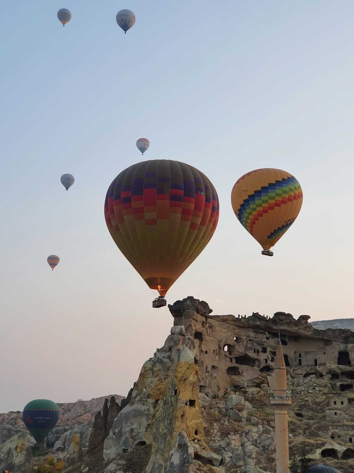 Une série de montgolfières dans le ciel, au-dessus d’un paysage rocheux parsemé de formations rocheuses anciennes et d’une structure dotée de multiples ouvertures.