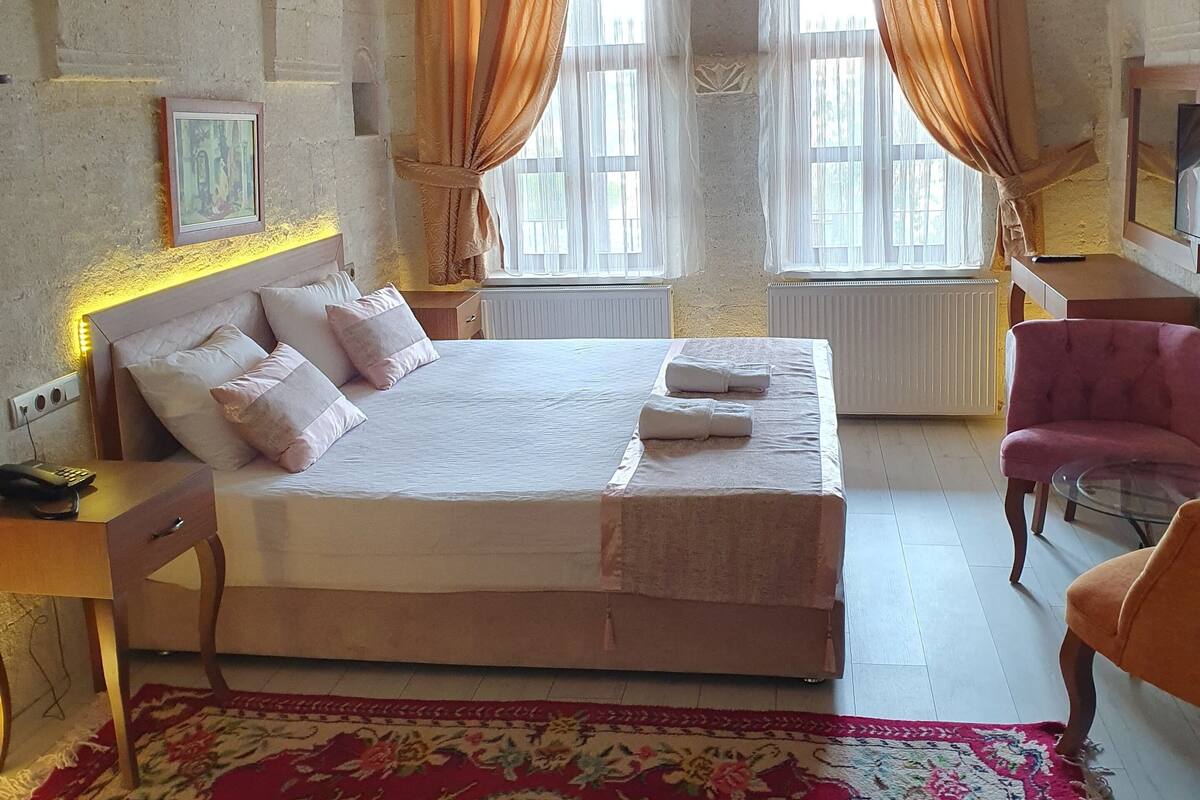 A spacious bedroom with a large bed, two windows with curtains, a radiator, a desk with a chair, and a patterned rug.
