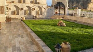 A courtyard with a stone building, a grassy area, and a large rock formation in the background. - Mesken Cave Suites (Avanos)