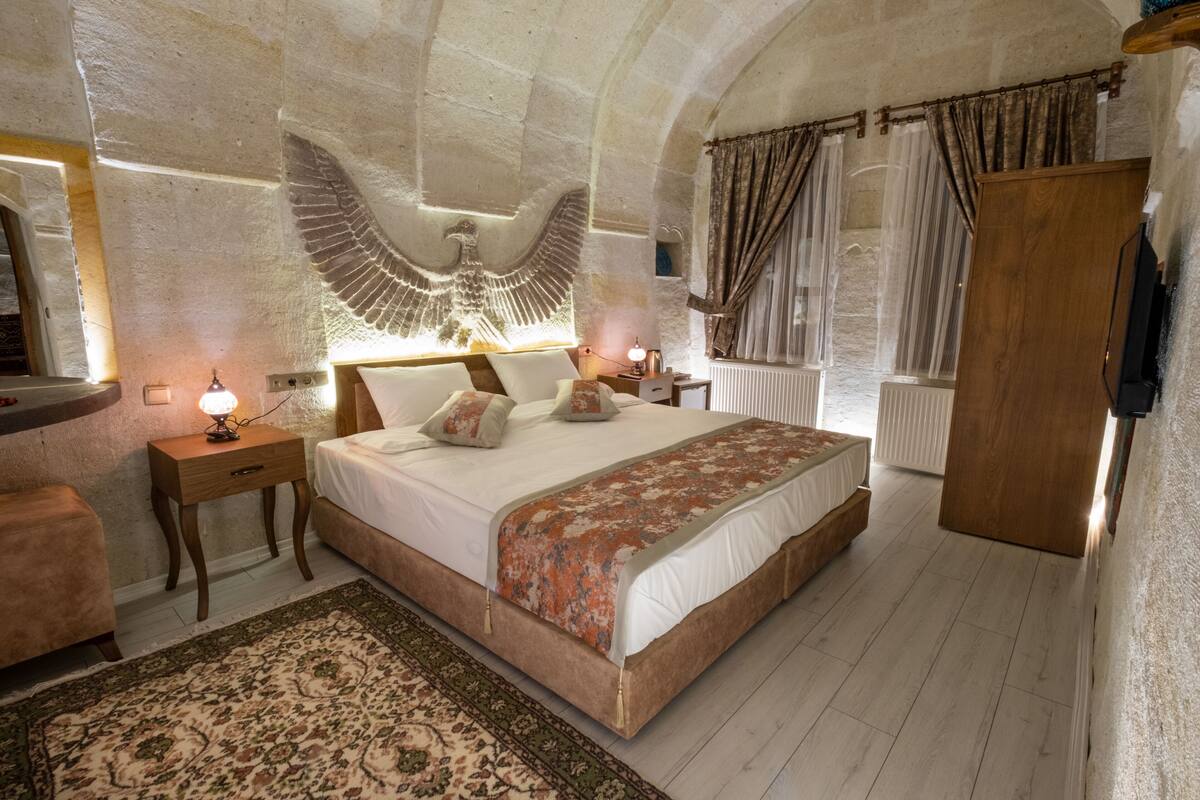 A bedroom with a large bed, a wooden nightstand, a bedside table with a lamp, a decorative rug, and a stone wall with an eagle carving.