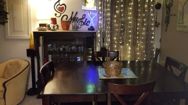 Un área de comedor con una mesa de madera, sillas, un muro decorativo con un corazón y una nota musical, y una guirnalda de luces.
