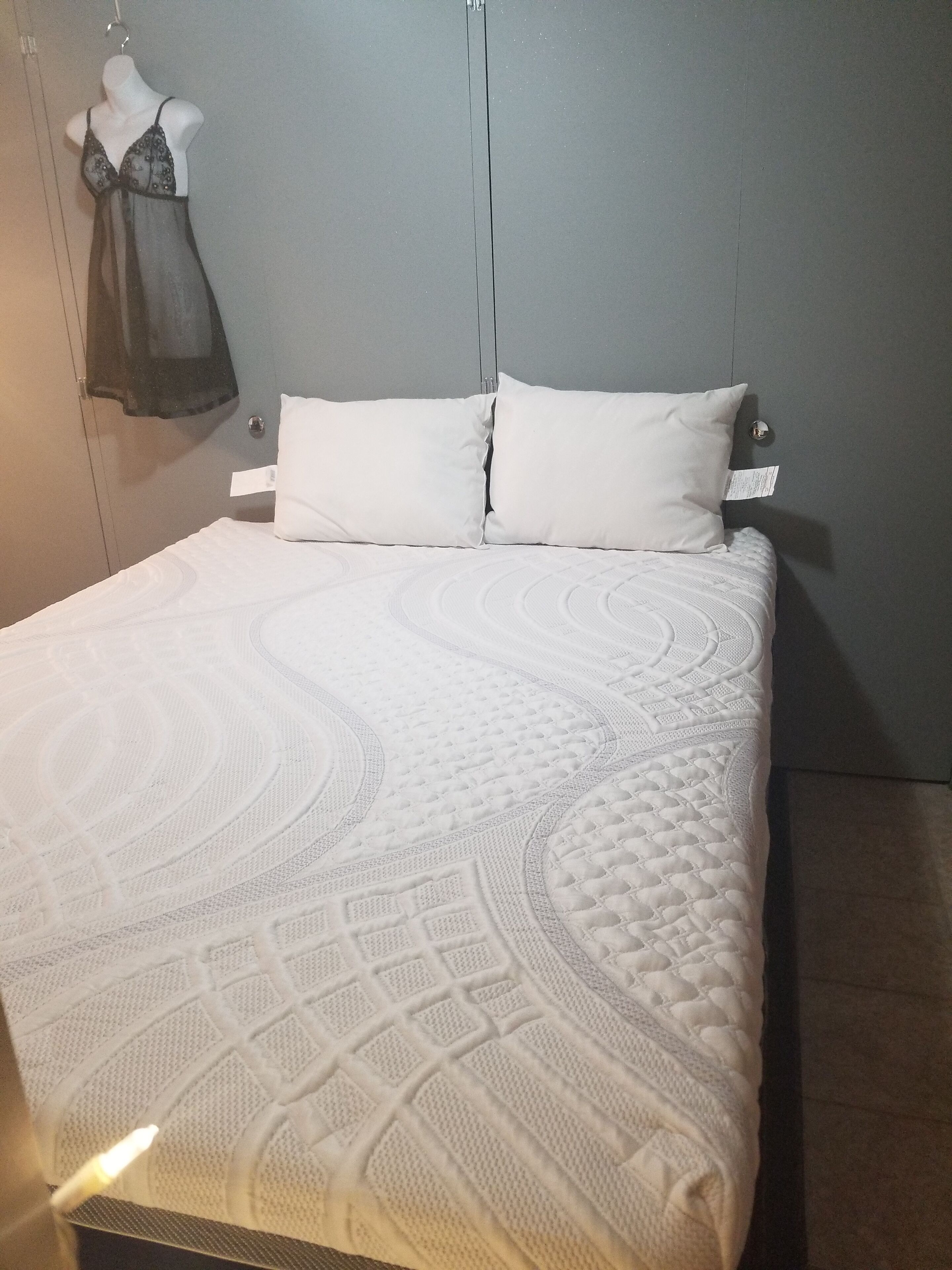 A bed with a white quilt and two pillows, a white dress on a mannequin, and a grey wall.