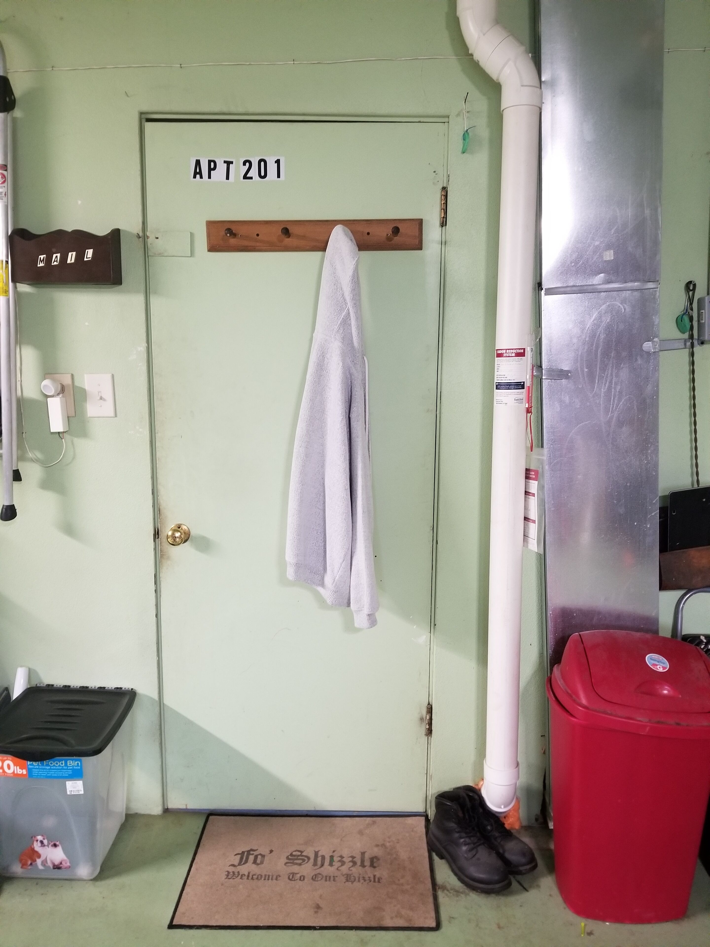 Une porte portant l’inscription « APT201 », avec une serviette suspendue à un crochet, un récipient pour la nourriture des animaux, une poubelle rouge et une paire de chaussures posées sur le sol.