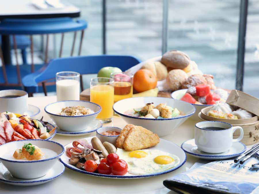 Une table dressée pour le petit-déjeuner, avec différents plats, notamment des œufs, des saucisses et des fruits, et une vue sur l’eau.