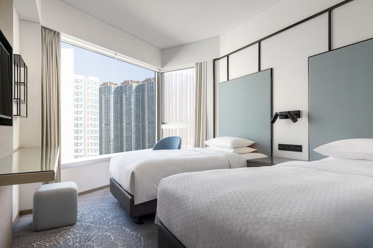 A hotel room with two beds, a large window with a city view, a desk with a chair, and a small ottoman.